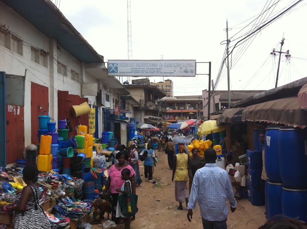 Shoppers and traders on a busy market street in Kumasi, stalls stacked with buckets and sandals
