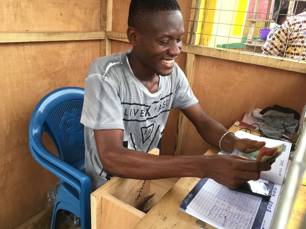 A young man smiling as he counts cash at his wooden mobile-money kiosk, a ledger open beside him