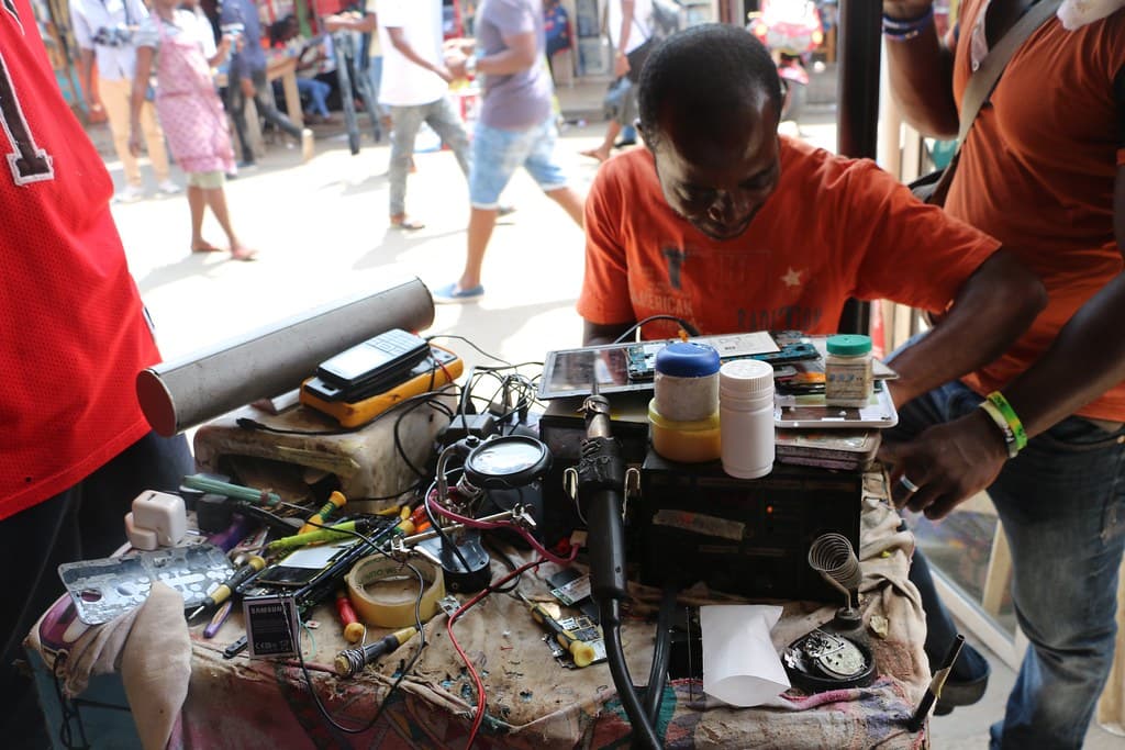 A repairer in an orange shirt works on phone circuit boards at a street workbench in Accra