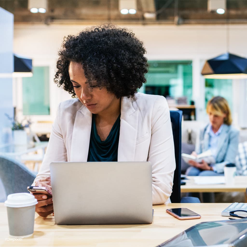 A woman in a pale blazer checks her phone beside a laptop at a shared office desk